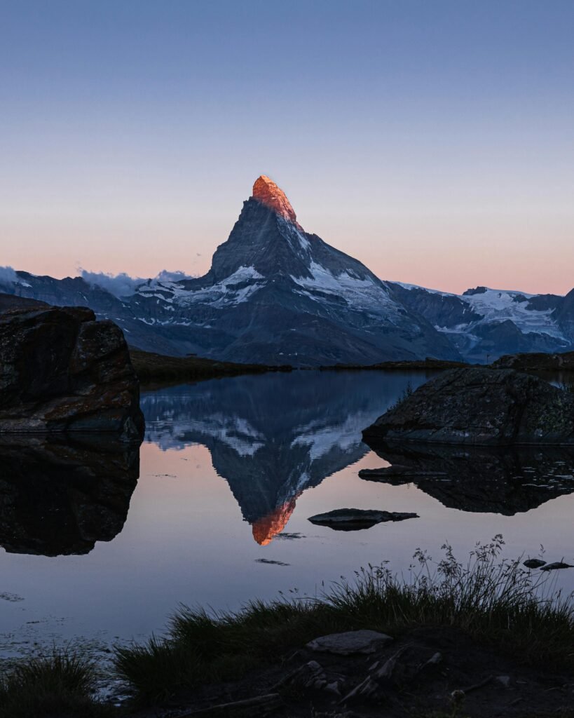 Matterhorn kalnas su saulės apšviesta viršūne ir atspindžiu skaidriame ežere. Paveikslėlis simbolizuoja galia ir asmeninį augimą, susijusius su saviverte, asmeniniu tobulėjimu, sąmoningumu ir lyderyste, skatinant moters atsiskleidimą. / Matterhorn mountain with a sunlit peak and reflection in a clear lake. The image symbolizes power and personal growth related to self-esteem, personal development, mindfulness, and leadership, promoting a woman's self-discovery.