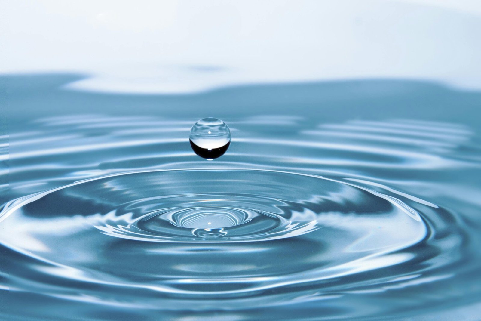 Vandens lašas mėlyname raibuliuojančiame vandenyje, simbolizuojantis puikią savijautą. / Water droplet in blue rippling water, symbolizing excellent well-being.